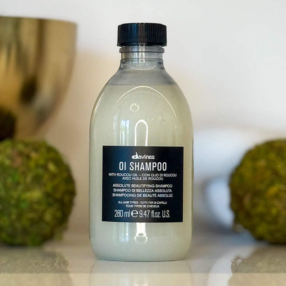 Bottle of Davines OI Shampoo with a black cap on a white surface with green fruits in the background.