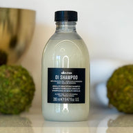 Bottle of Davines OI Shampoo with a black cap on a white surface with green fruits in the background.